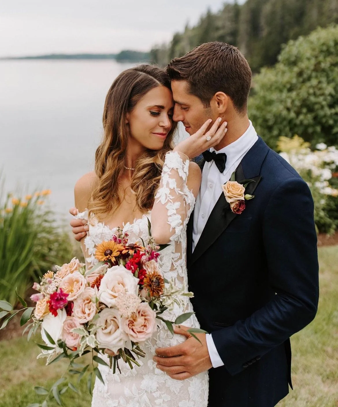 A newlywed couple shares a kiss outdoors by a lake, with the bride holding a bouquet of flowers and the groom wearing a tuxedo.