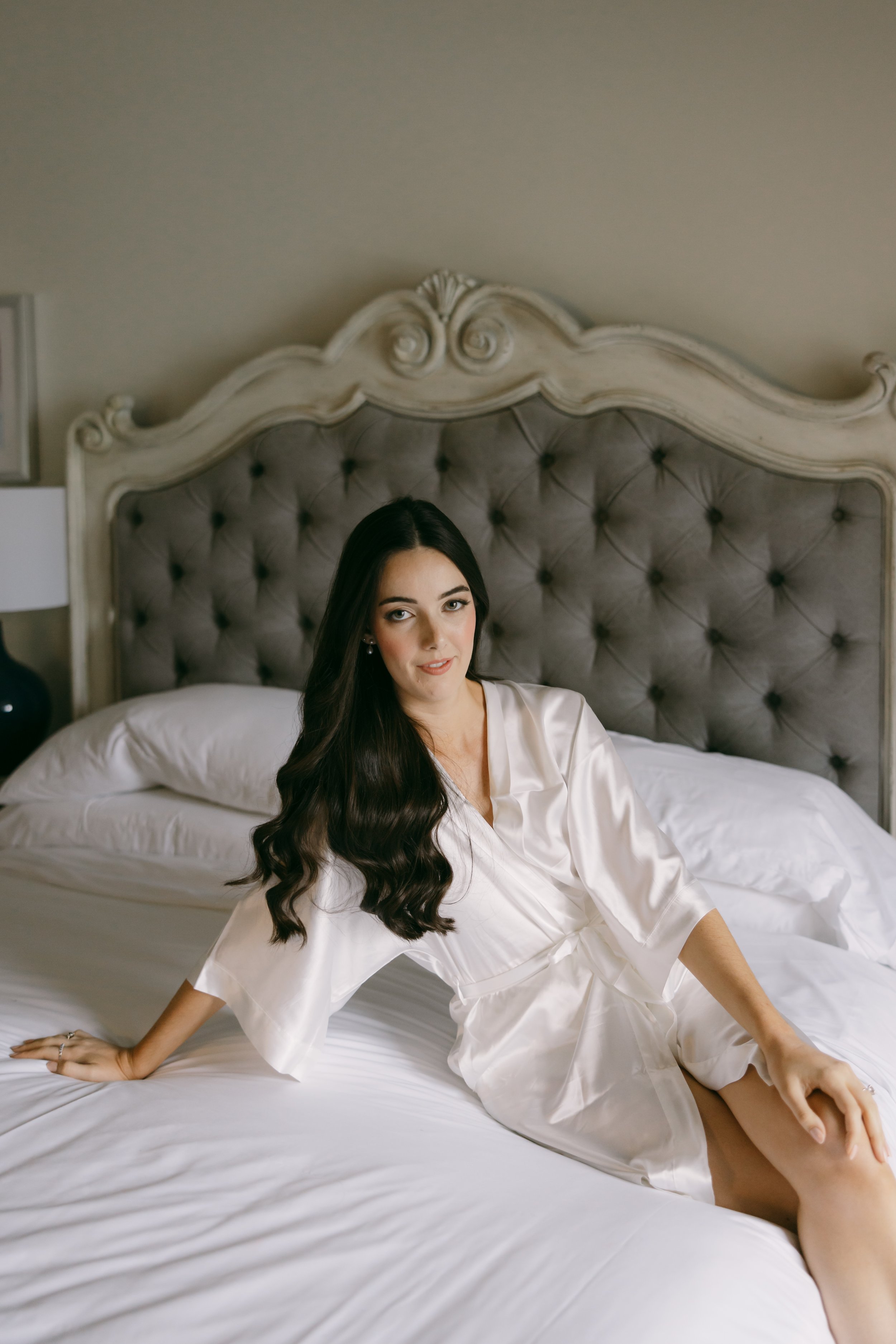 A woman with long dark hair sitting on a bed with a tufted headboard, wearing a silky white robe.