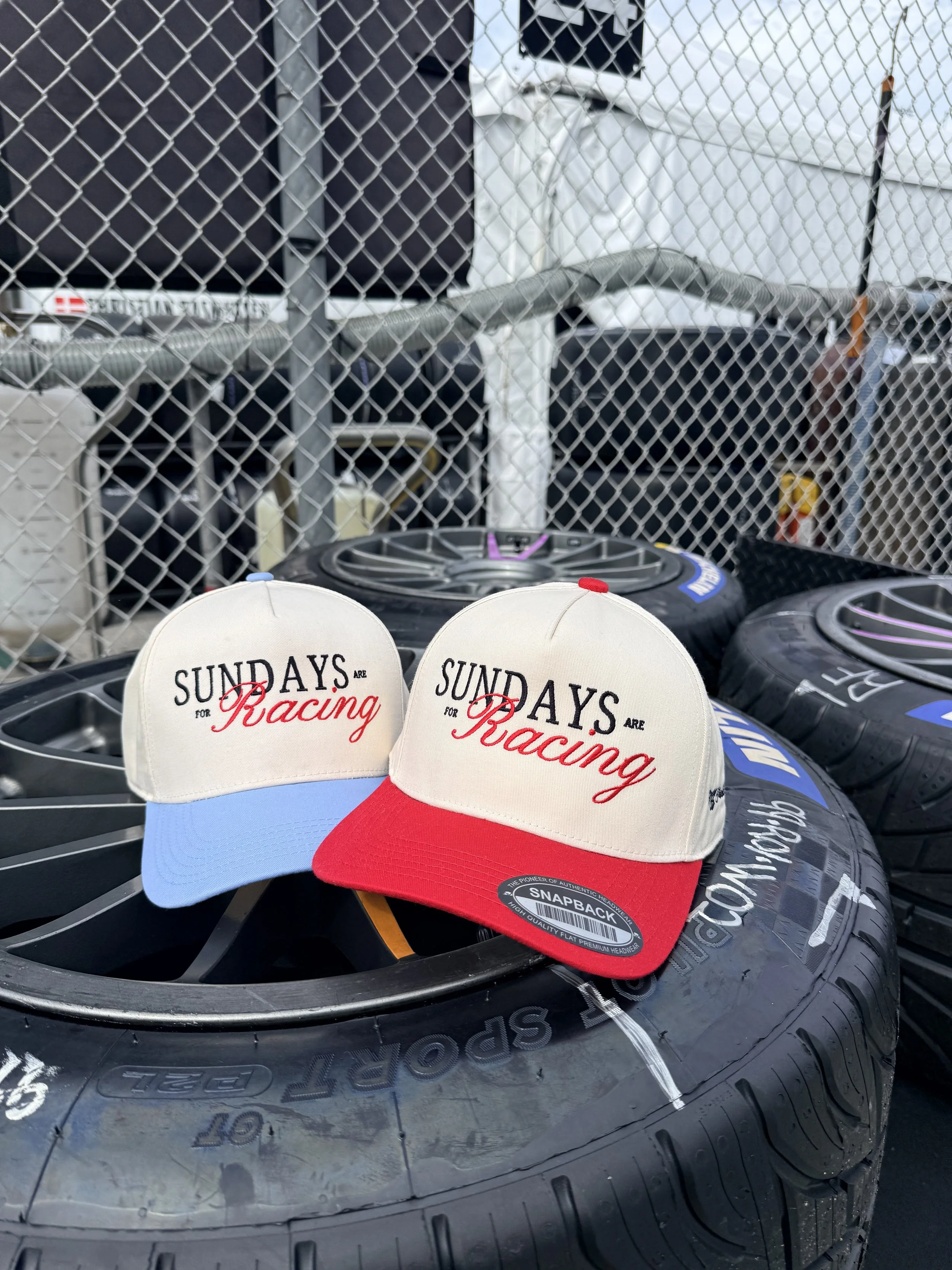 Two racing hats with the words 'Sundays are for Racing' placed on a tire next to other tires behind a chain-link fence.