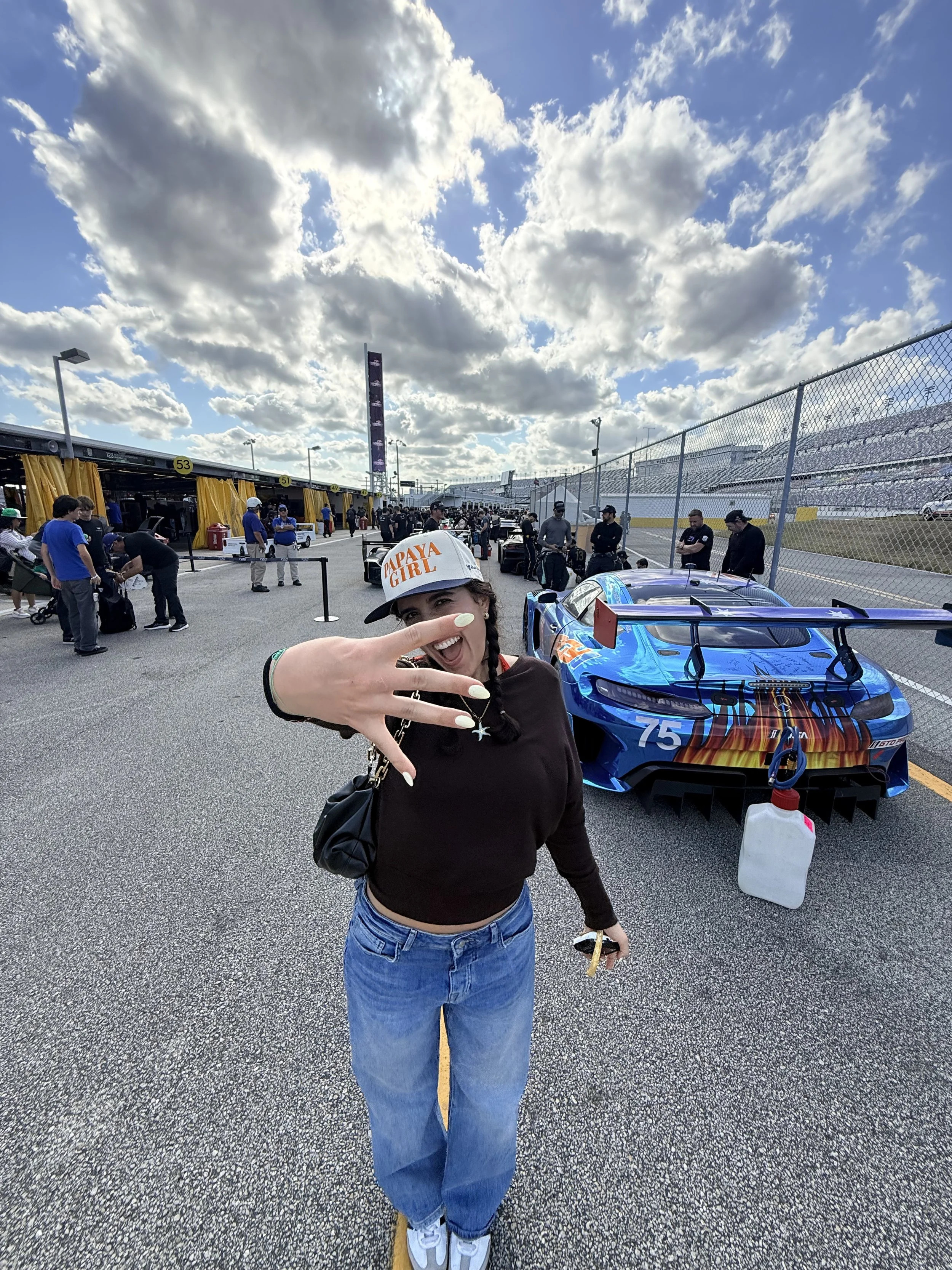 A woman wearing a Papaya Girl cap and a black top, showing a peace sign, standing on a race track with a blue race car in the background, surrounded by people and race facilities.
