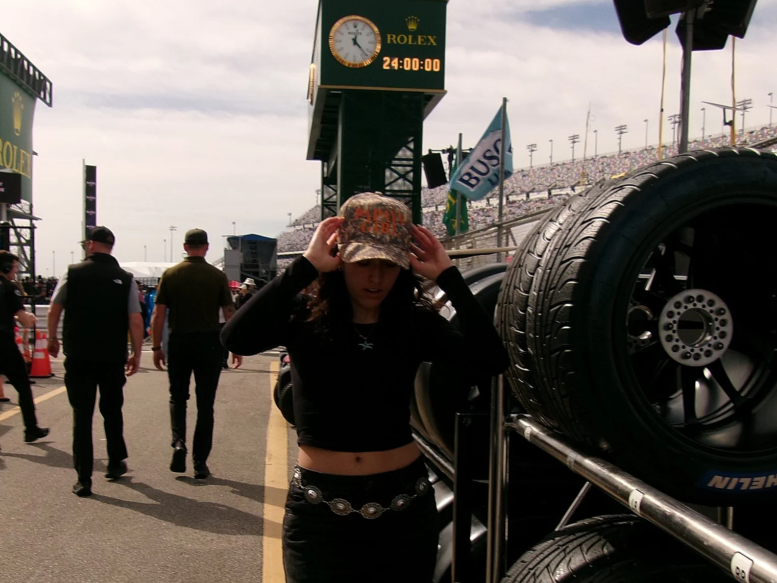 A young woman wearing a camouflage cap and black outfit adjusting her cap at a racing event, with people walking and a large tire display in the background, along with a Rolex clock on a scoreboard.
