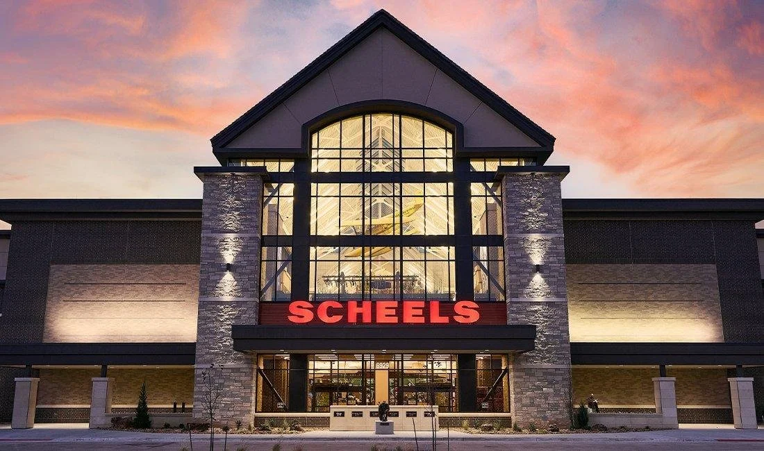 Cityscape with the entrance to Scheels store at sunset, featuring a large glass facade, stone pillars, and red Scheels sign.