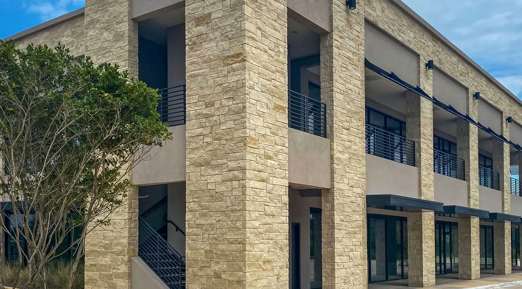 Modern multi-story building with stone and concrete exterior, multiple balconies with metal railings, large glass windows, and a tree in the foreground.