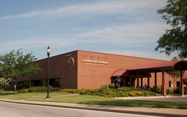 The Indian Health Care building, a brick structure with a red roof canopy, is located on a grassy area with trees and a lamp post in front.
