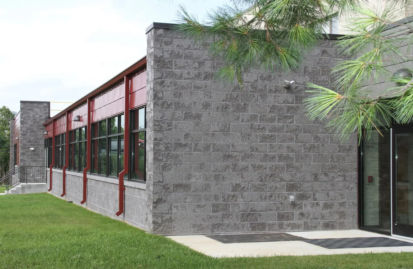 The exterior of a modern building with gray concrete block walls, large windows with red frames, and a glass door. There is a green lawn, a concrete pathway, and partial view of pine tree branches.
