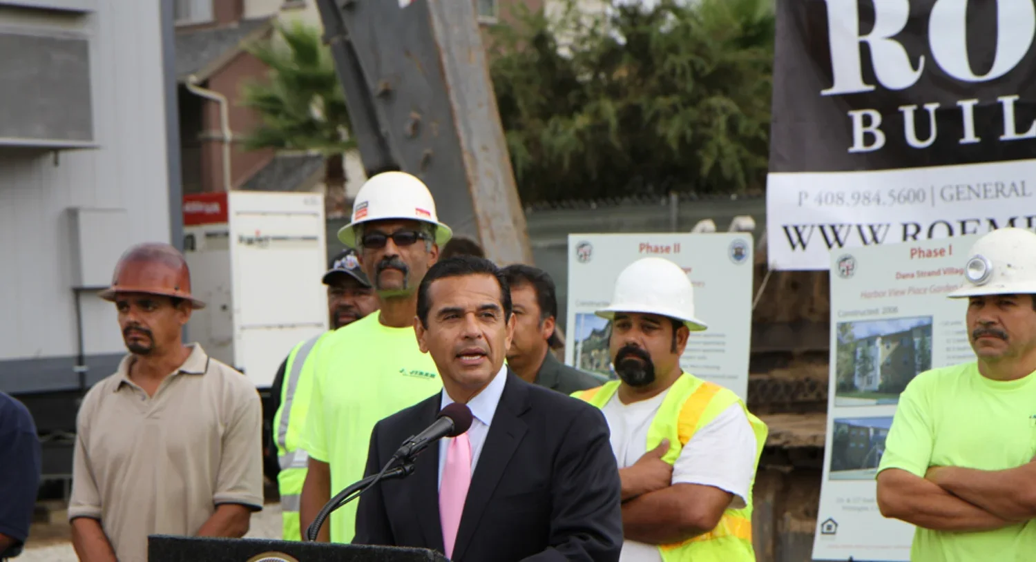 A man in a suit and pink tie speaking at a podium, with construction workers in safety vests and helmets behind him during a press conference at a construction site.