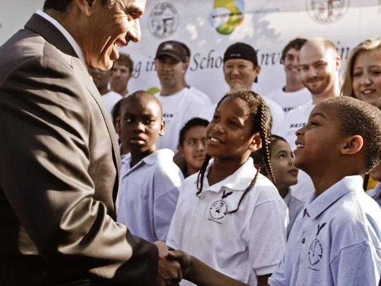 A man in a suit shaking hands with a young girl in a white polo shirt, surrounded by smiling children and adults at an outdoor event.