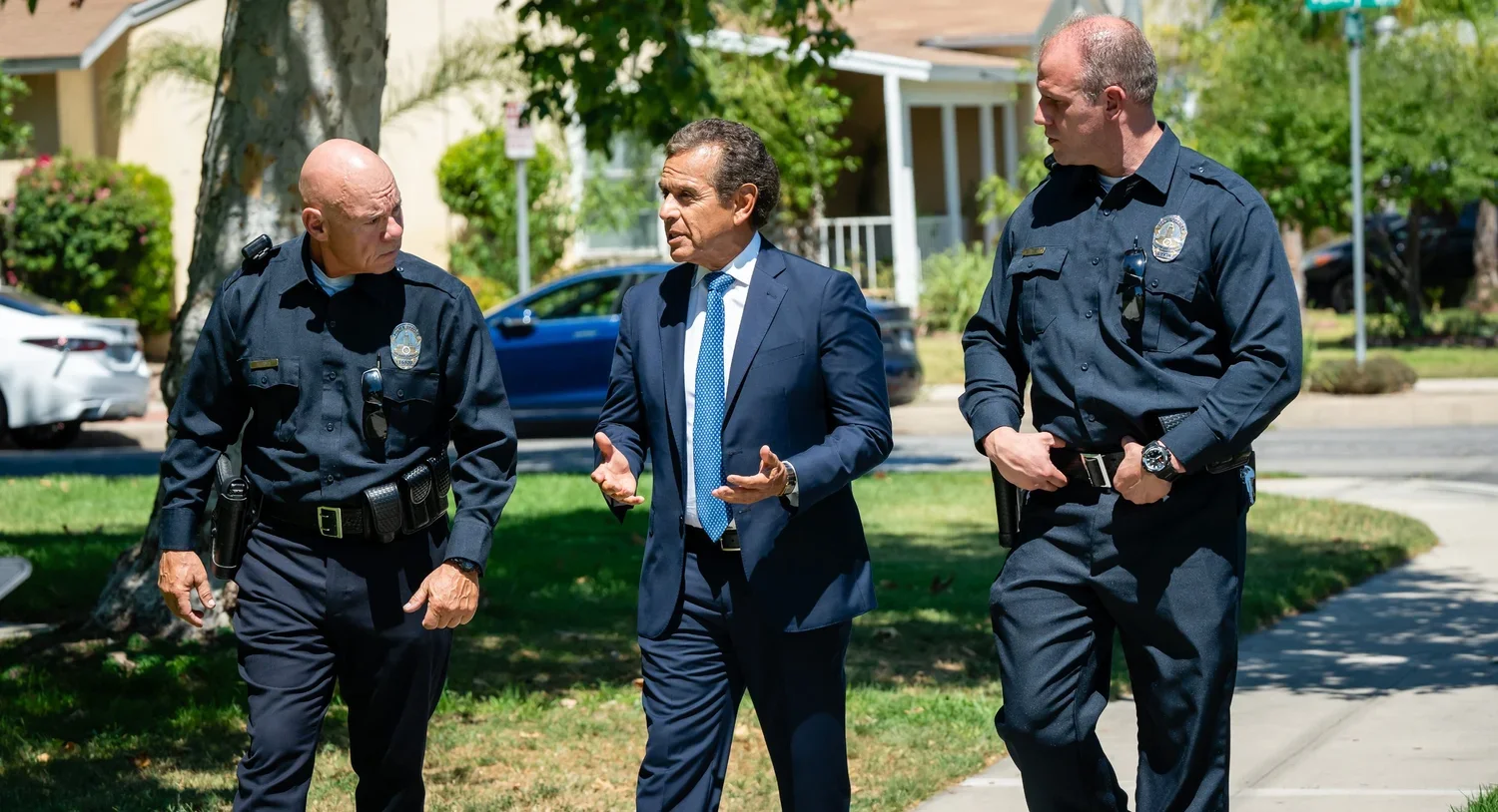 Three men walking outdoors, two police officers in uniform and one man in a suit, engaged in conversation, with houses and parked cars in the background.