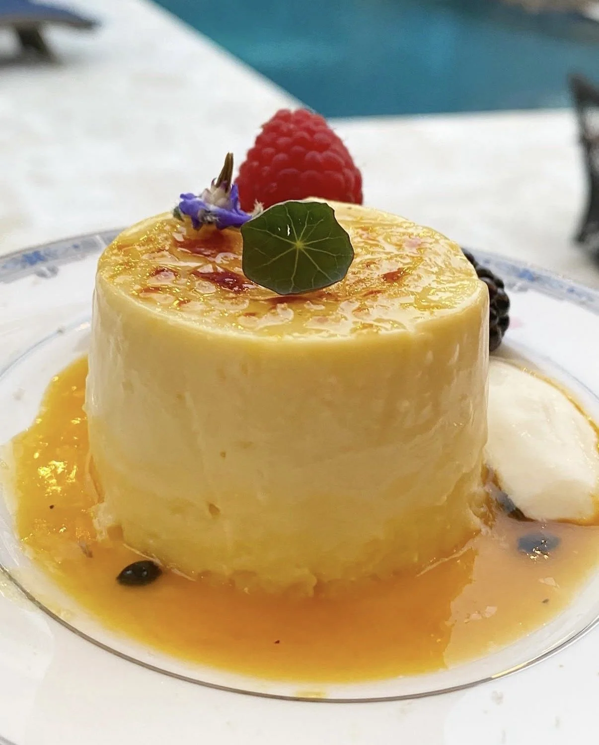 A dessert with a glazed yellow cake topped with a raspberry, edible flower, and a green leaf, served on a white plate with sauce and berries.