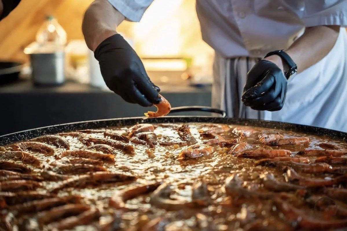Chef frying shrimp in a large skillet, wearing black gloves and white apron.