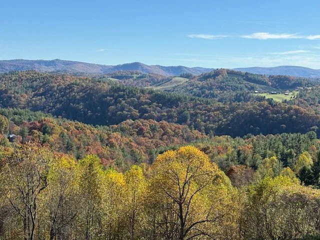 Landscape of rolling hills with colorful autumn trees, distant mountains, and a blue sky with some clouds.