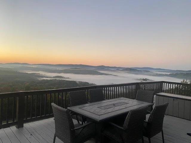 Outdoor patio with a dining table and six chairs overlooking a foggy valley at sunrise or sunset.