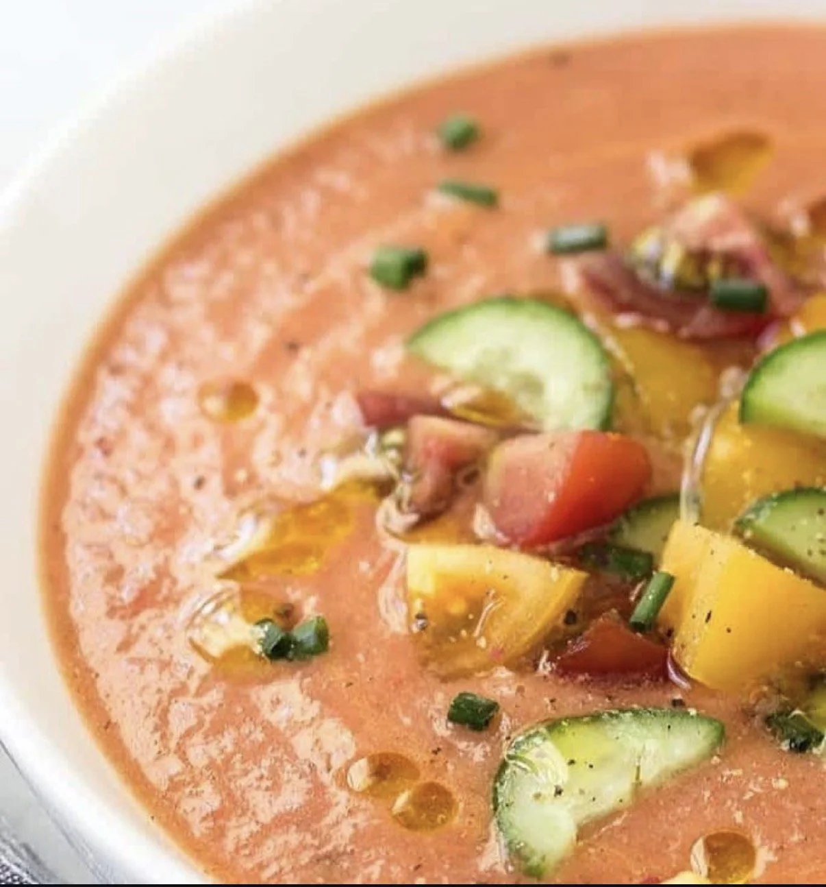 Close-up of a bowl of tomato-based gazpacho soup garnished with cucumber slices, chopped chives, and small pieces of chopped vegetables.