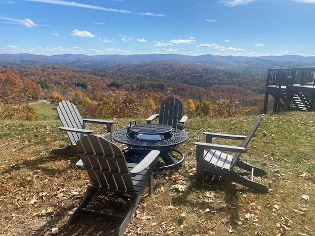 Outdoor patio with four black Adirondack chairs arranged around a fire pit on a grassy hilltop, overlooking a mountain landscape with colorful fall foliage and a partly cloudy sky.