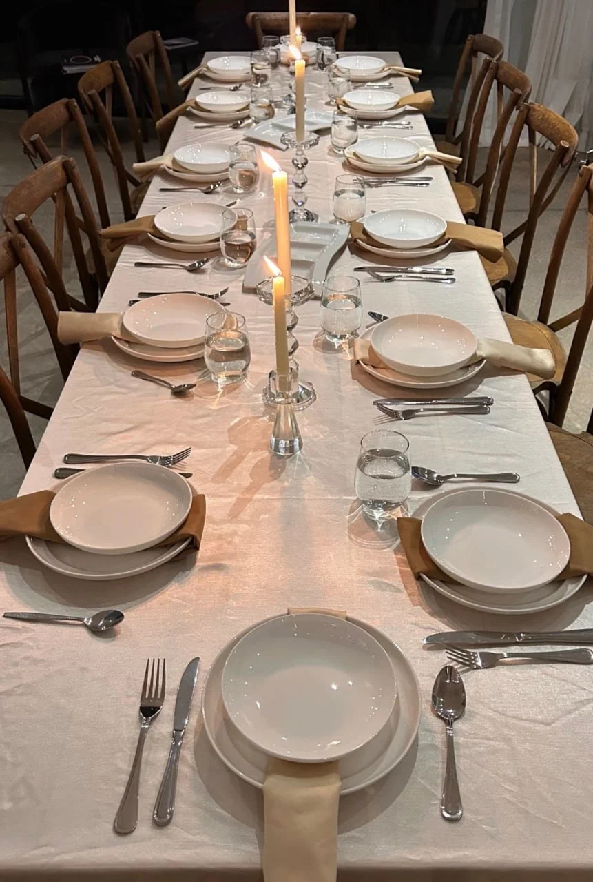A long dining table set for a formal dinner with white tablecloth, multiple place settings including white plates, bowls, glasses of water, and silverware, centered by tall lit candles in candlesticks.