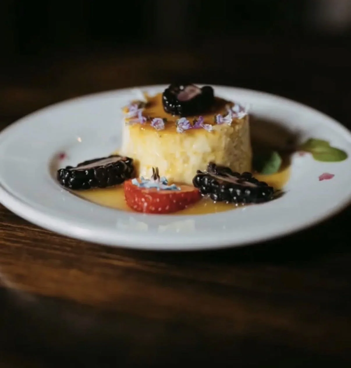A dessert on a white plate featuring a flan garnished with blackberries, strawberries, and edible flowers, with syrup drizzled over it.
