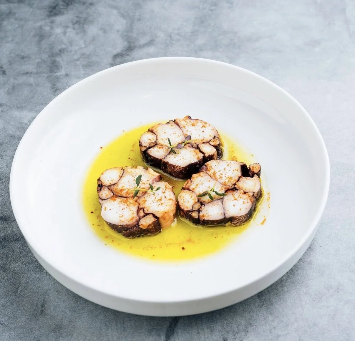 Three pieces of octopus on a white plate, drizzled with olive oil and garnished with herbs.