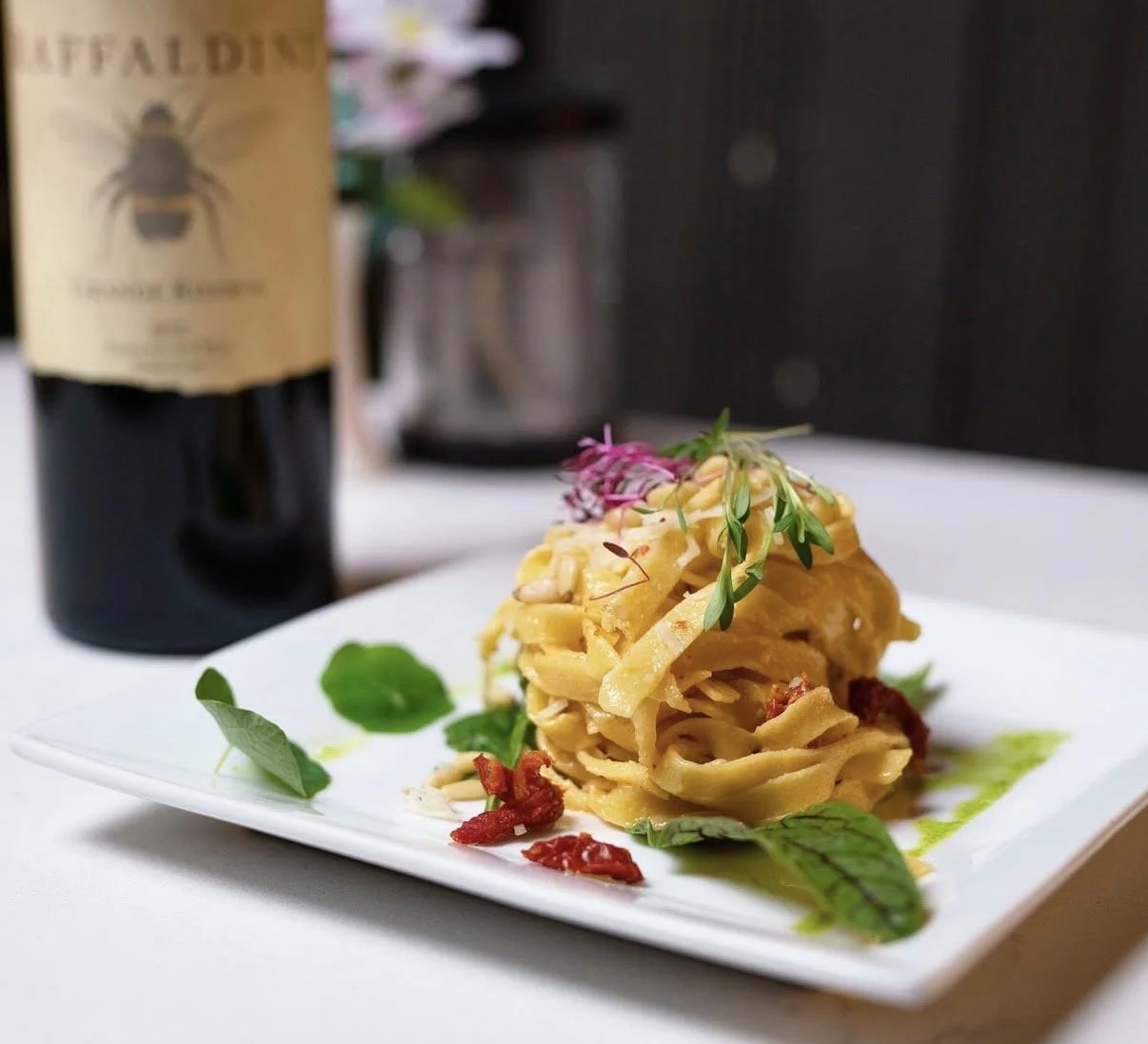Plate of pasta with vegetables and herbs, garnished with microgreens, served with a glass of red wine in the background.