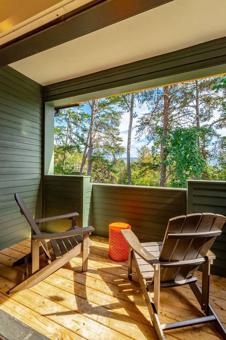 A balcony with wooden flooring, two Adirondack chairs, an orange side table, surrounded by green trees, overlooking a forested area.