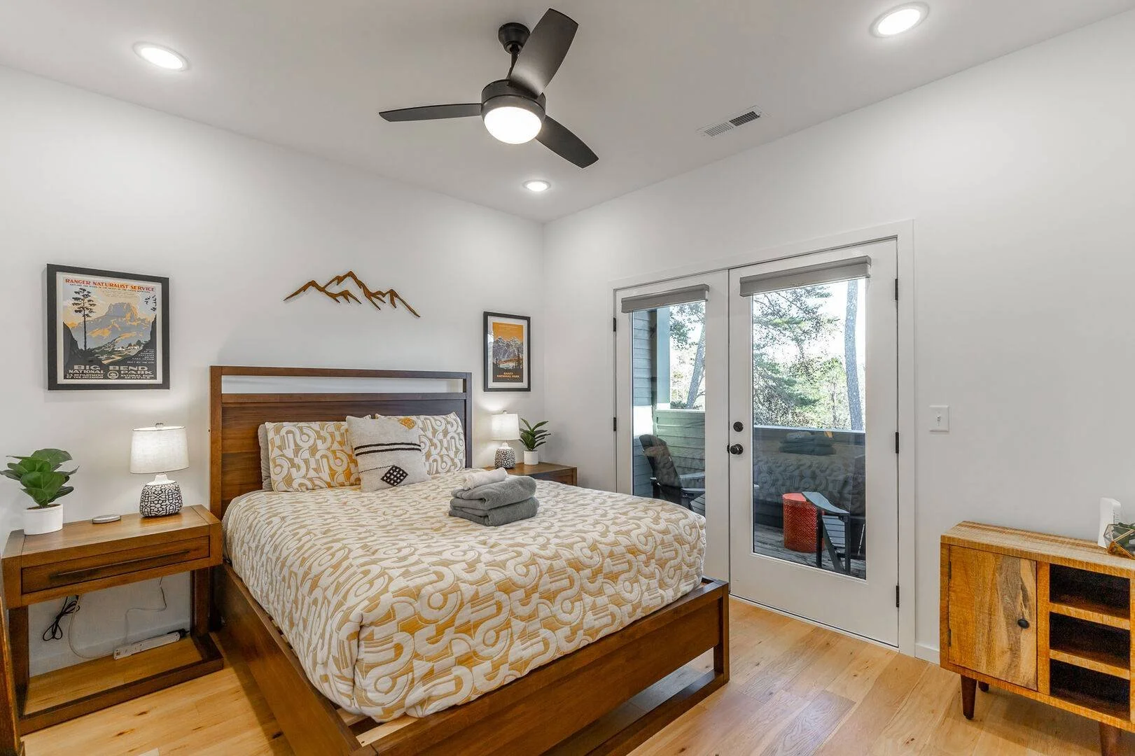 A queen bedroom with two nightstands with lamps on either side of the bed, decorative objects and plants. A glass door leads to a balcony with outdoor furniture and trees visible outside.