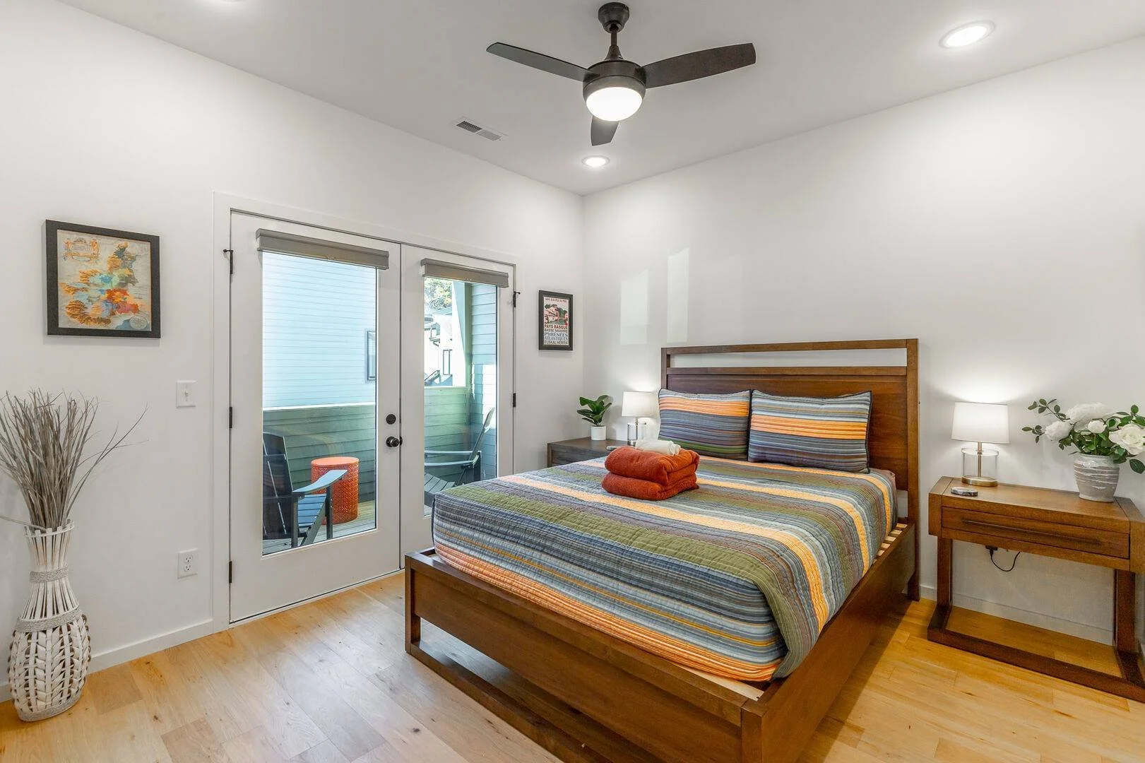 A queen bedroom with two bedside tables with lamps and a plant, wooden floor, ceiling fan, and glass door leading to an outdoor balcony with chairs and decor.