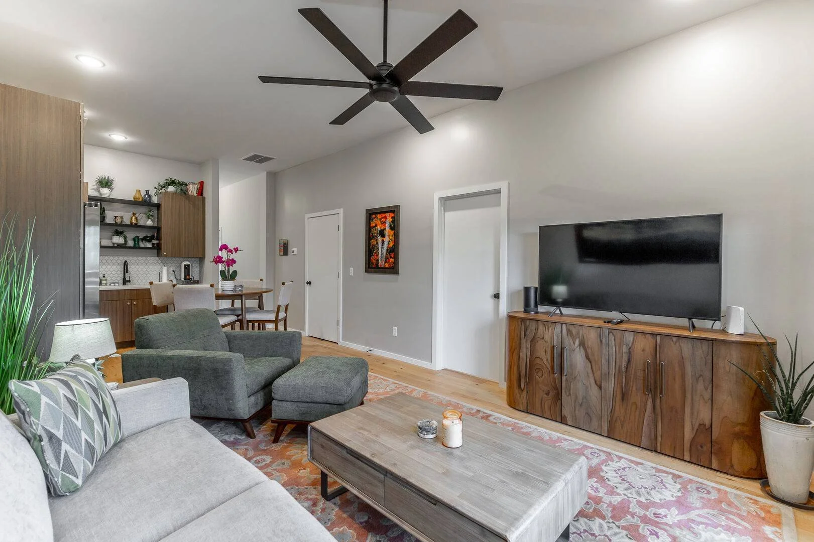 Living room with a sofa, armchair, and ottoman, a TV stand with a flat-screen TV, a potted plant, a coffee table with candles, adjacent to a dining area with a round table and chairs, and an open kitchen.