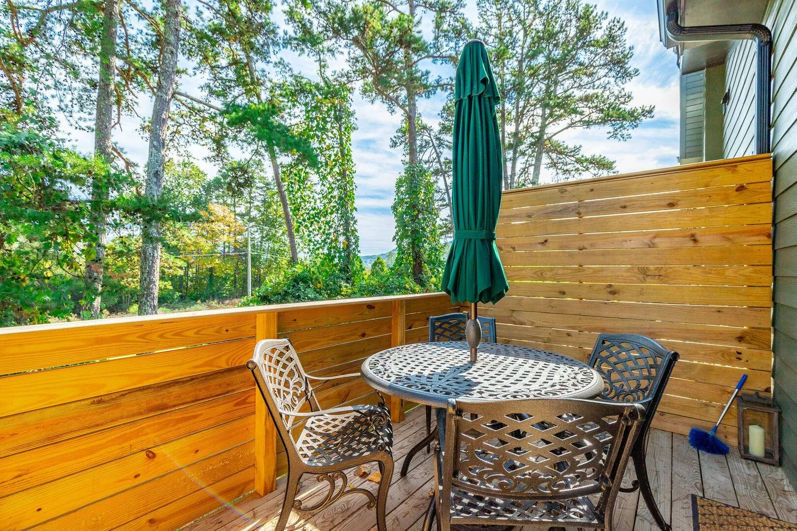 An outdoor balcony with a round table, four chairs, umbrella, and a privacy wall, overlooking trees and sky.