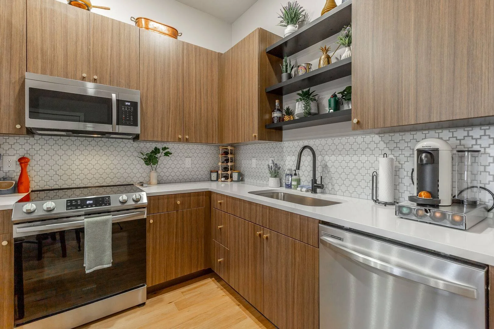 Kitchen with cabinets, tile backsplash, microwave, stove, and stainless steel refrigerator. There are plants and decorative items on open shelves and counter, with wooden flooring.