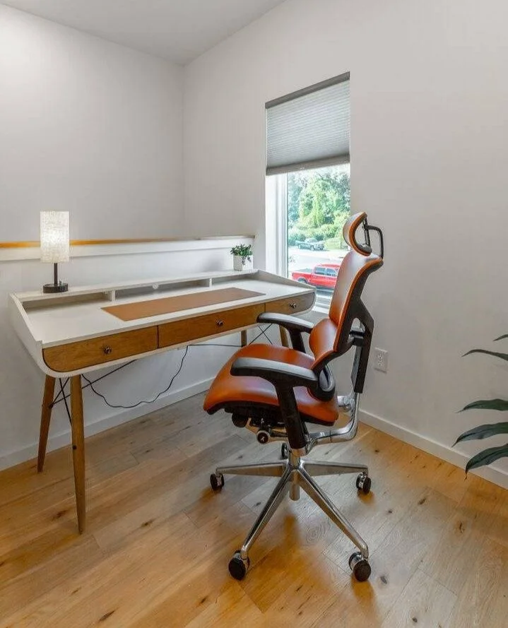 A modern, minimalist home office with a desk, a ergonomic chair, a small lamp, and a window showing green trees outside.