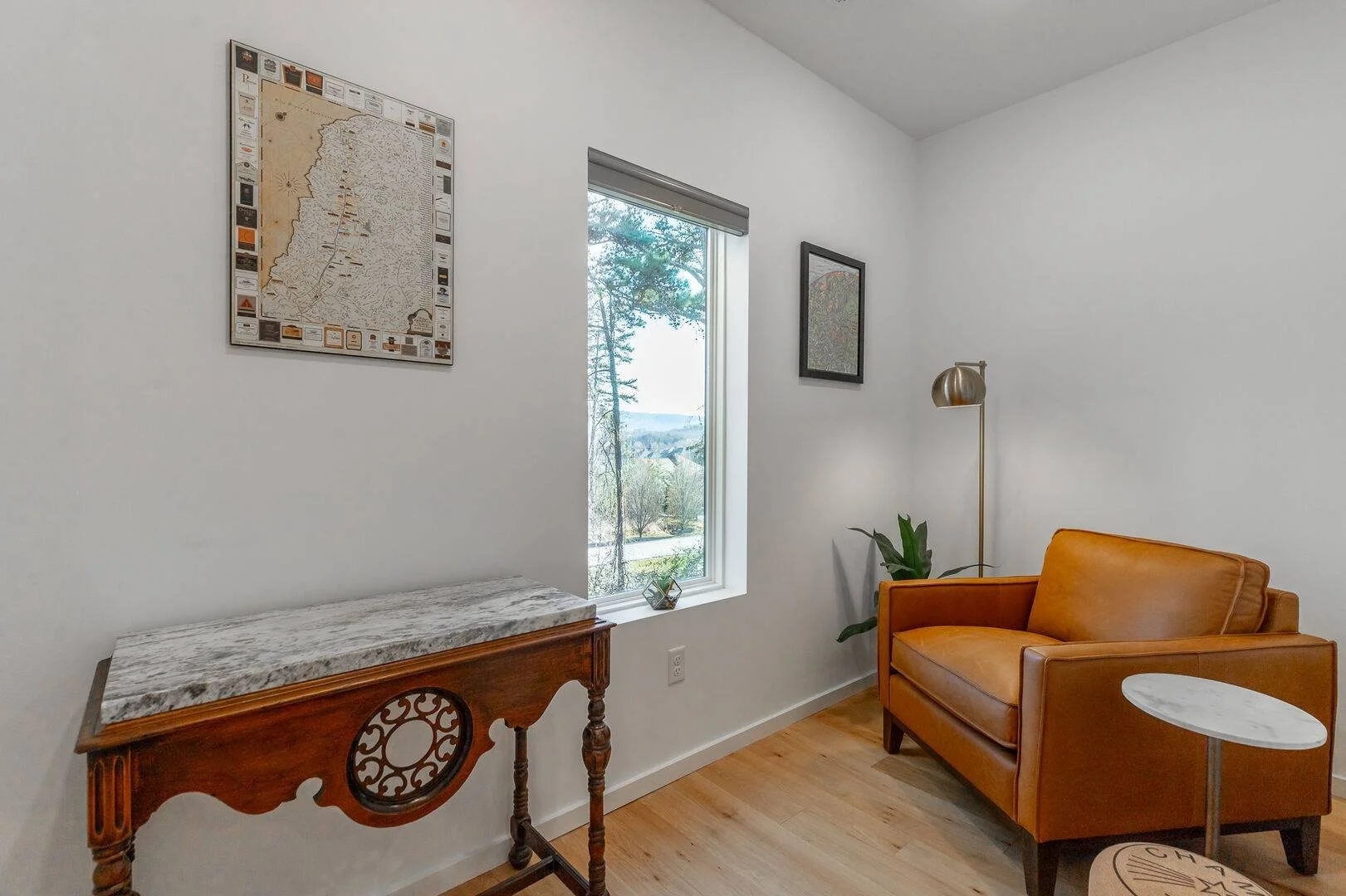 A sitting room with window and leather armchair, a tall standing lamp, a potted plant, a small round marble-top side table, and wooden flooring.