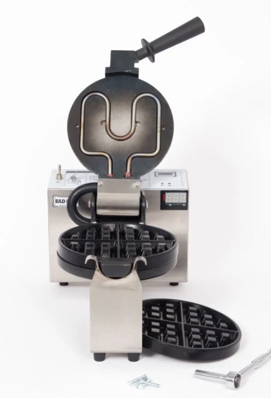 Commercial waffle iron with removable non-stick plates, closed lid, paddle spatula, and small tools beside it.