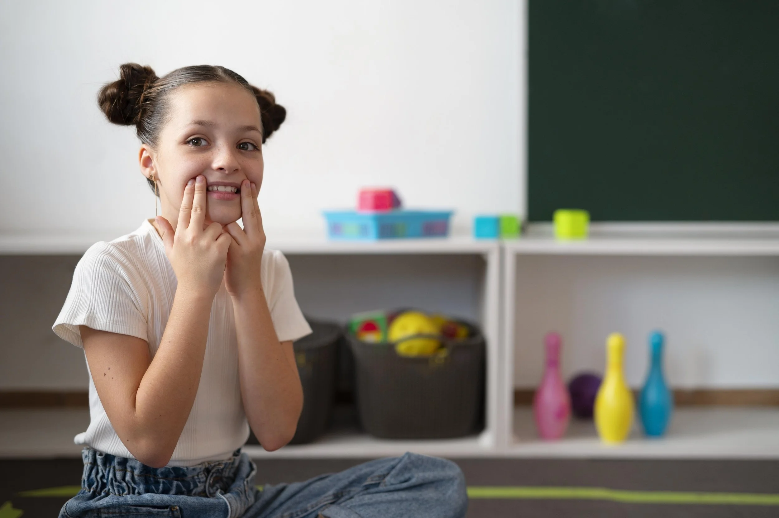 A young girl sitting cross-legged on the floor, smiling and holding her fingers on her cheeks, in a classroom with colorful toys and a chalkboard in the background.