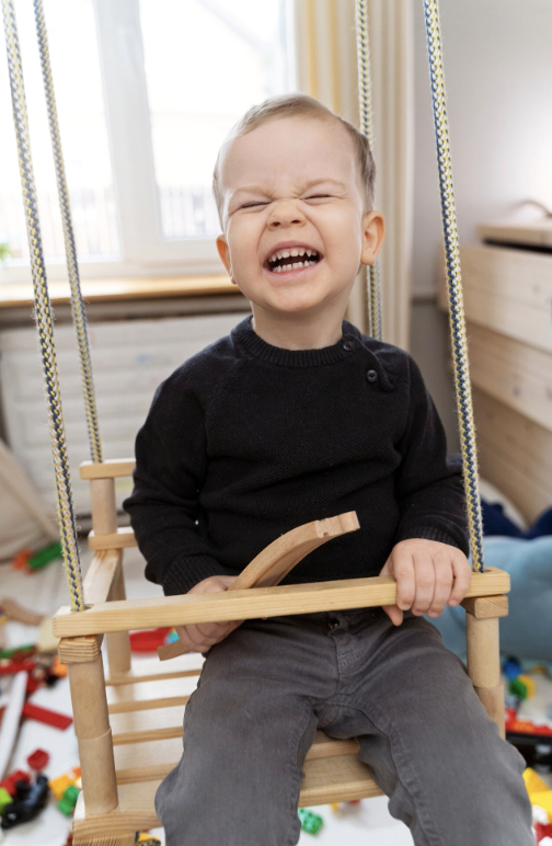 A young boy sitting on a wooden outdoor swing with a happy expression, surrounded by scattered toys.