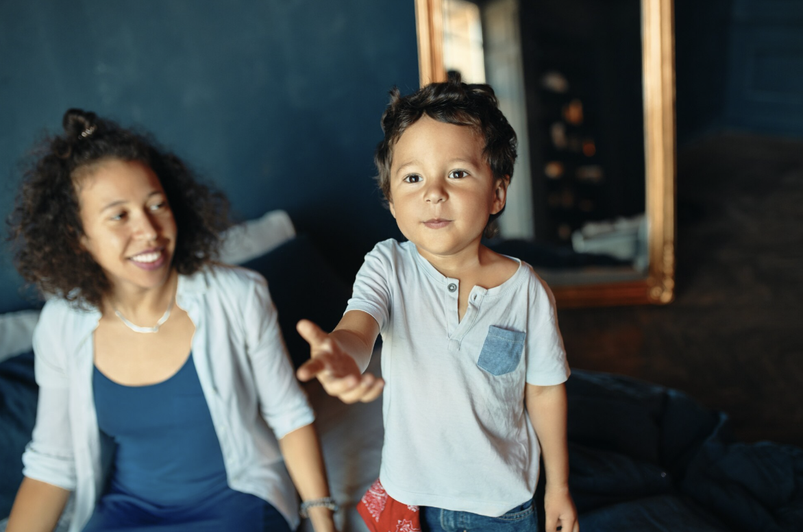 A young boy with dark curly hair reaching out his hand toward the camera, standing in a room with dark walls and a large mirror. A woman with curly hair and a bright smile, wearing a white shirt and blue dress, is sitting on the bed behind him, looking at him.