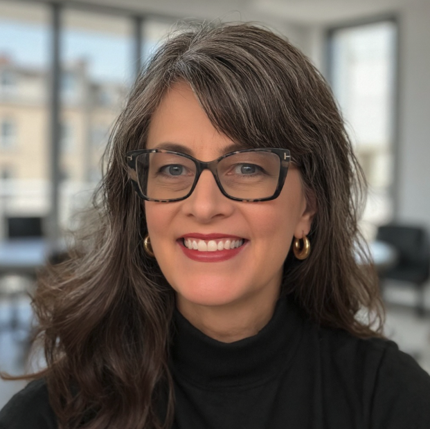 A smiling woman with shoulder-length brown hair and glasses, wearing a black turtleneck, in a bright, modern office with windows in the background.