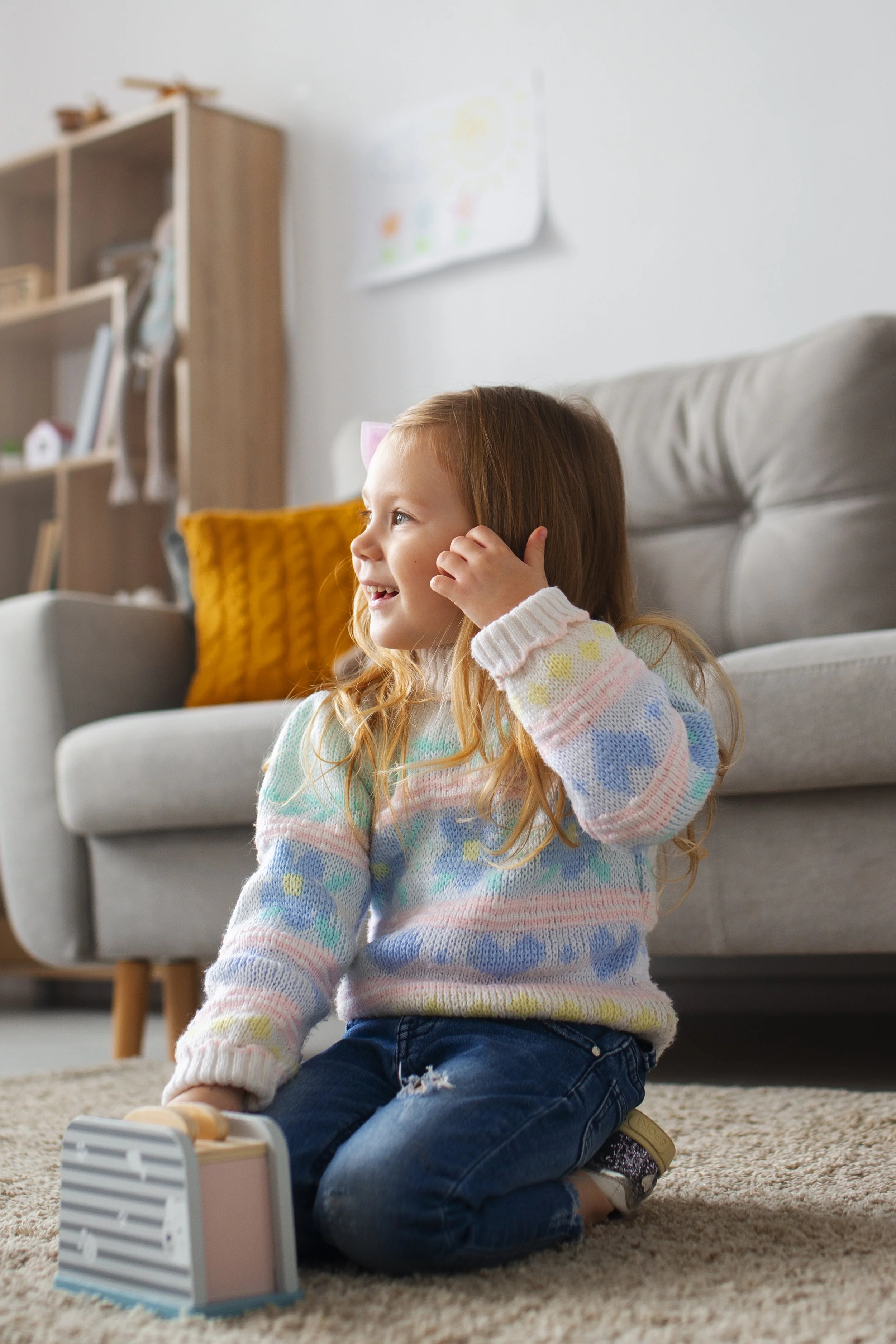 A young girl with red hair and a pink bow sitting on the carpeted floor, smiling and playing with a small wooden toy on a block in a living room.