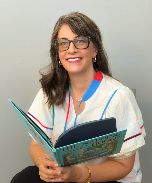 A woman with brown hair, glasses, and gold earrings smiling while holding an open children's book titled "The Napping House."