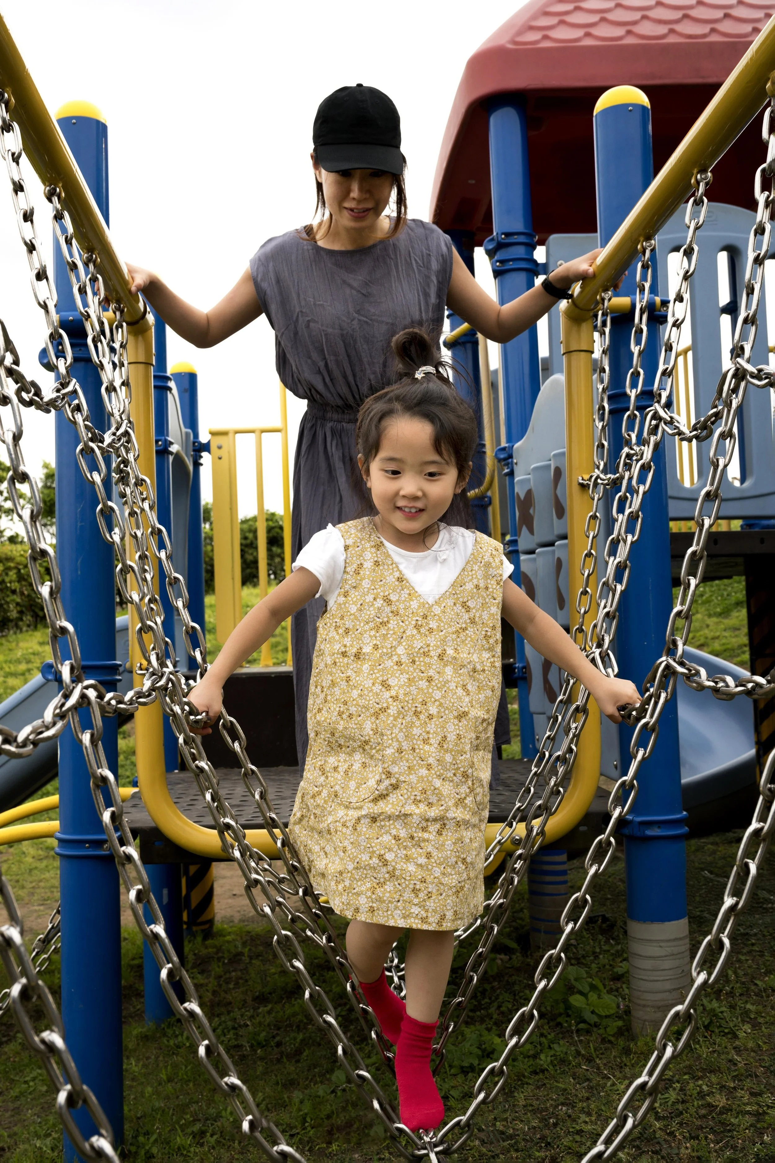 A woman and a young girl playing on a colorful playground structure outdoors. The girl is walking down a chain bridge, wearing a yellow dress with a floral pattern and red socks, while the woman watches behind her.