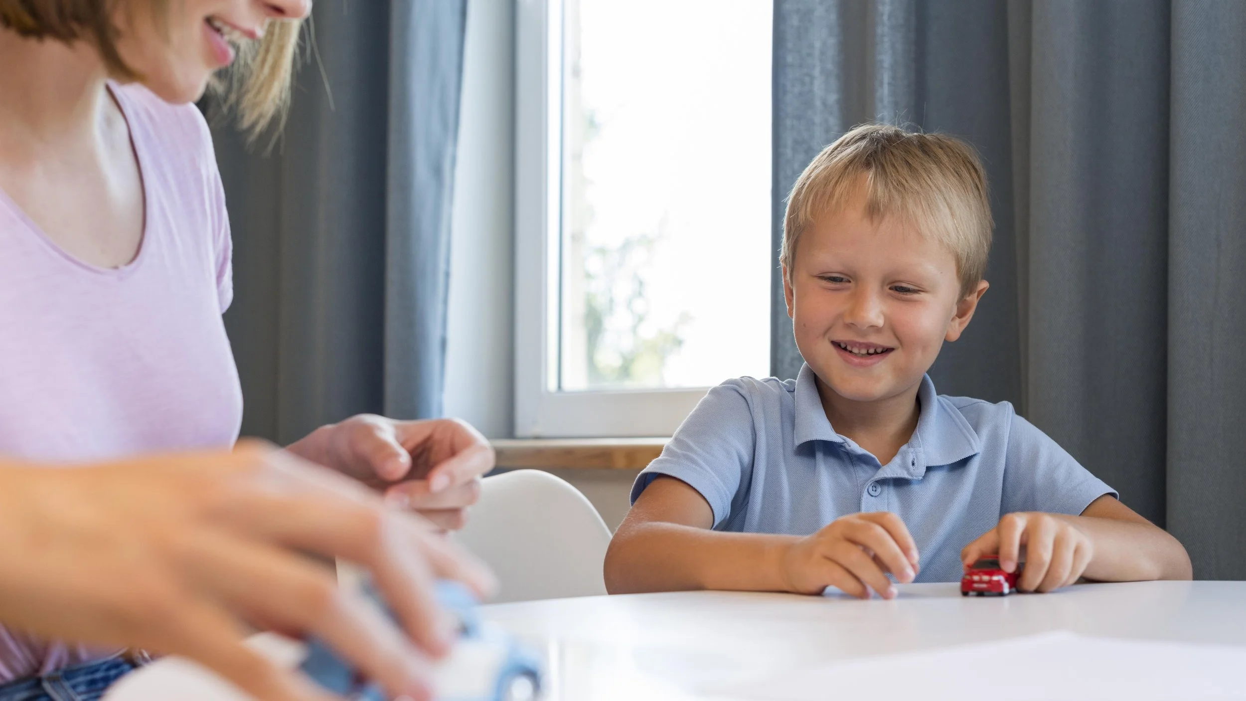 A young boy with blond hair smiling while playing with a small red toy car at a white table, with a woman partially visible on the left side, in a room with blue curtains and a window in the background.