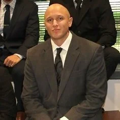 A man in a gray suit and black tie sitting in a formal setting, with other people partially visible in the background.