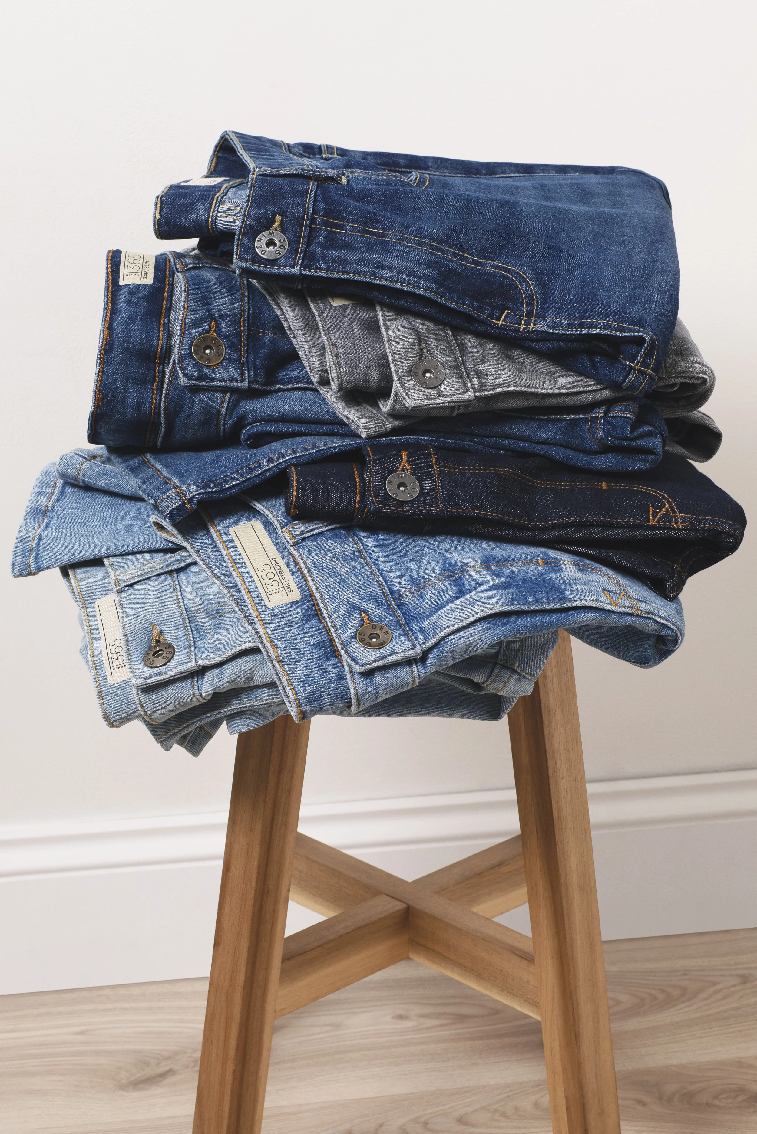 Stacked folded jeans of various shades on a wooden stool against a white wall.