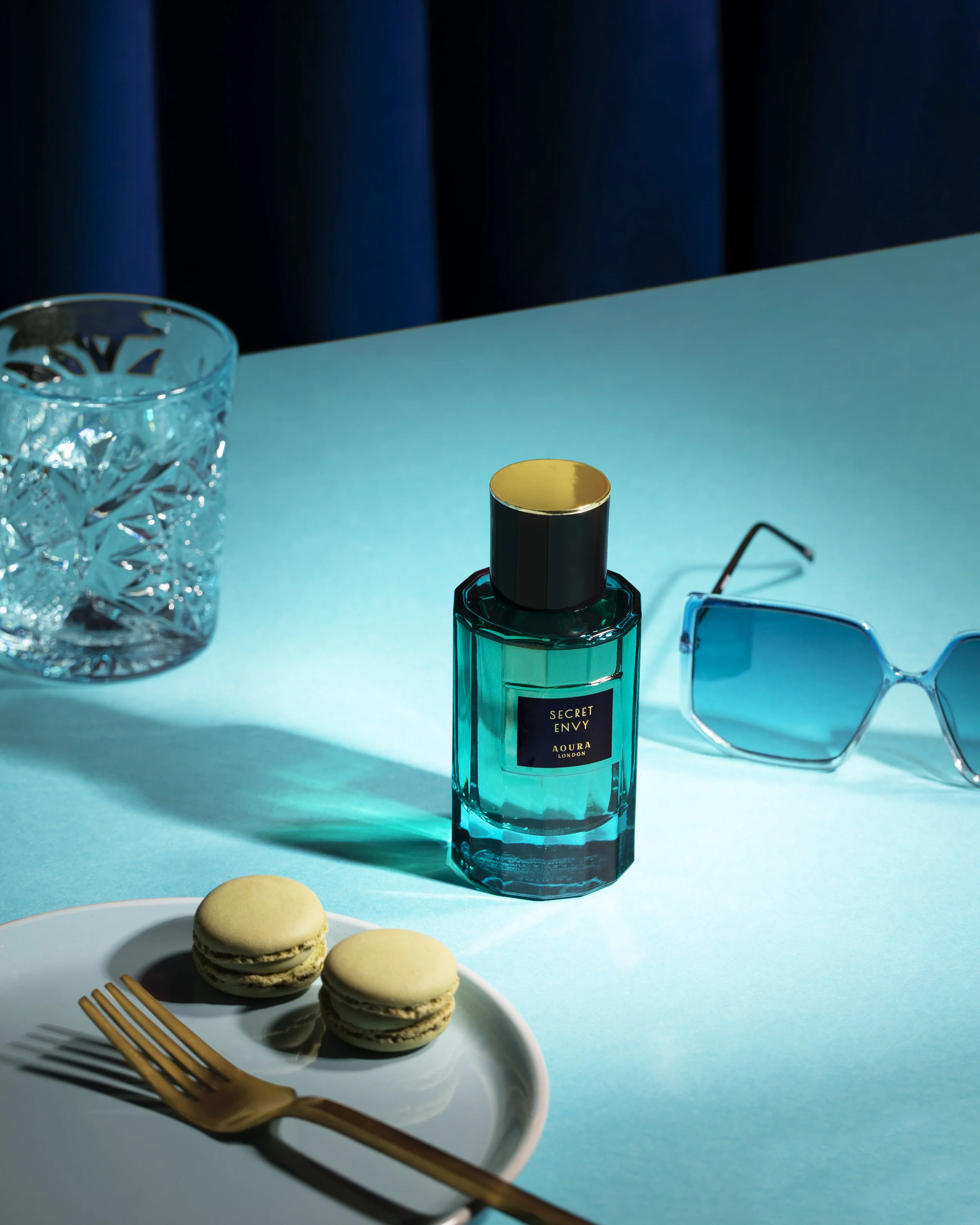 A glass perfume bottle labeled 'Secret Envy' by Aura London, a pair of large sunglasses with blue-tinted lenses, a white plate with two macarons and a gold fork, all on a teal surface with a dark curtain in the background.