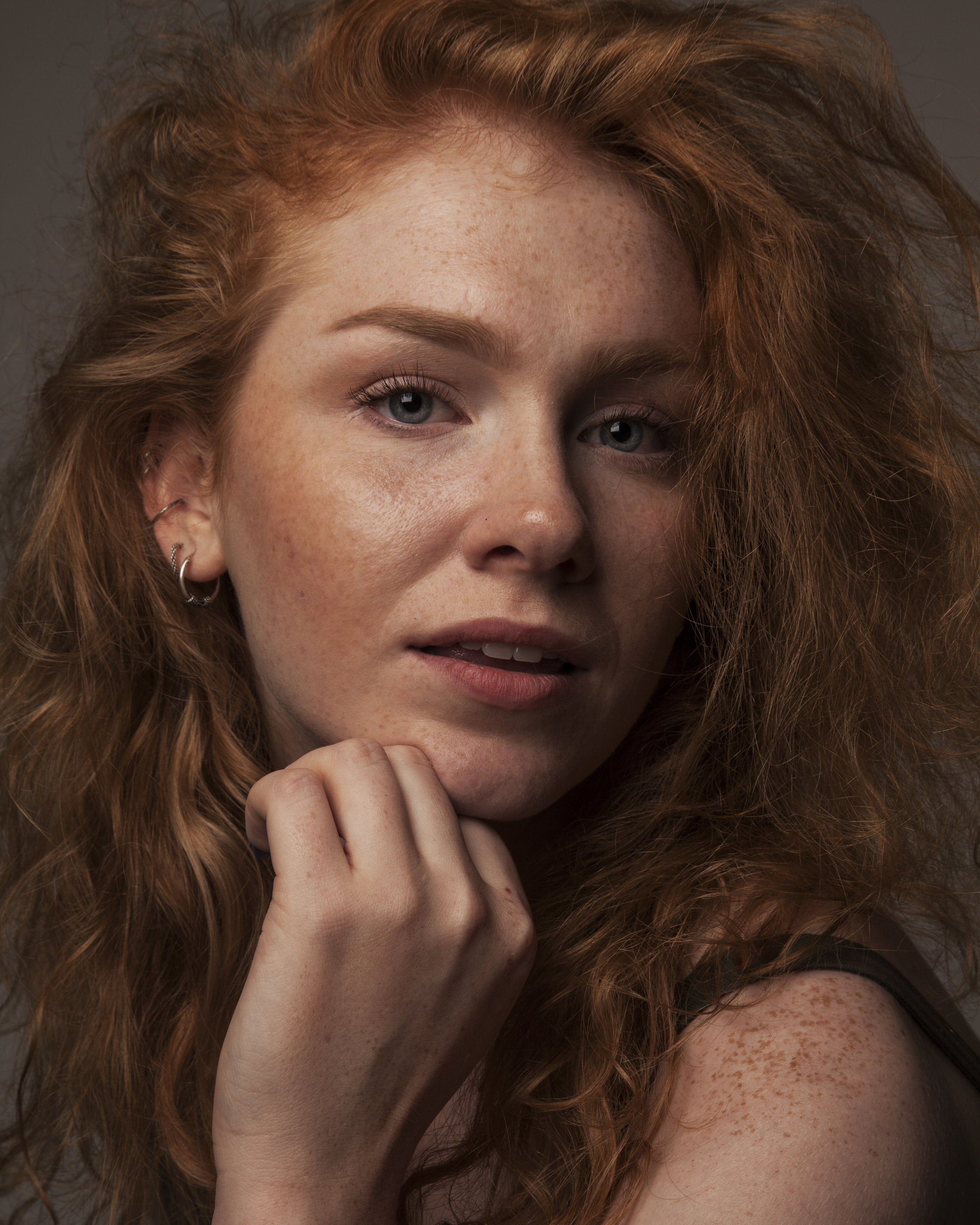 A close-up portrait of a young woman with red, wavy hair, light skin with freckles, and blue eyes, resting her chin on her hand and looking at the camera.