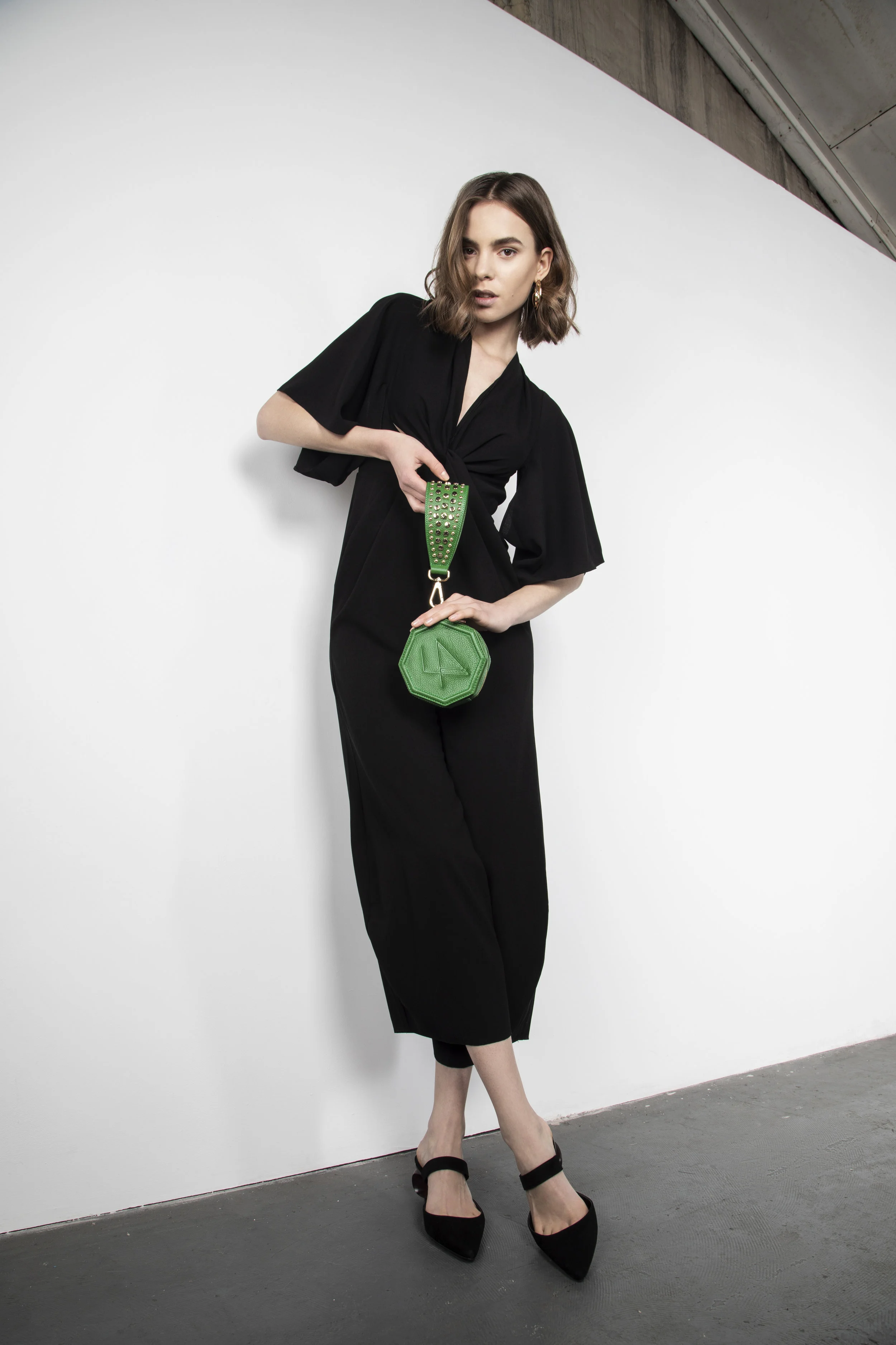 A woman in a black dress and black heels holding a green purse standing against a white background.