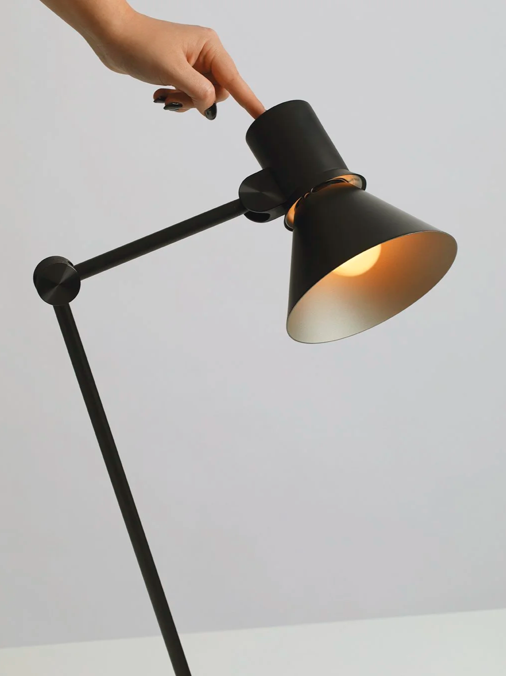 A hand with black painted nails adjusting a black adjustable desk lamp turned on, against a plain light gray background.