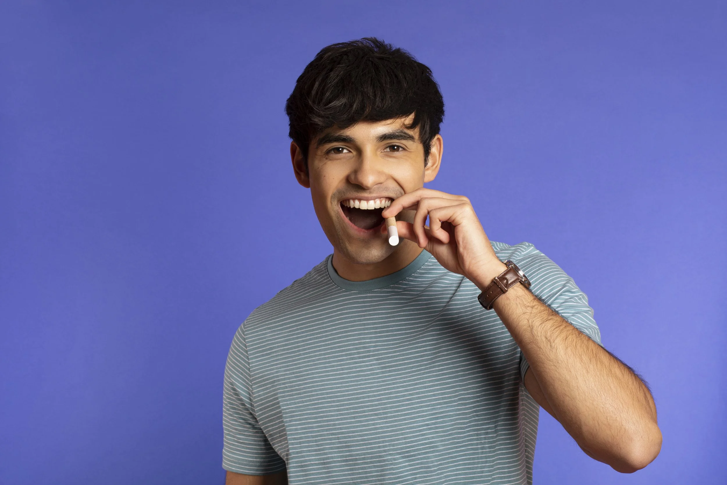 Young man with dark hair smiling and holding a cigarette near his mouth against a blue background.