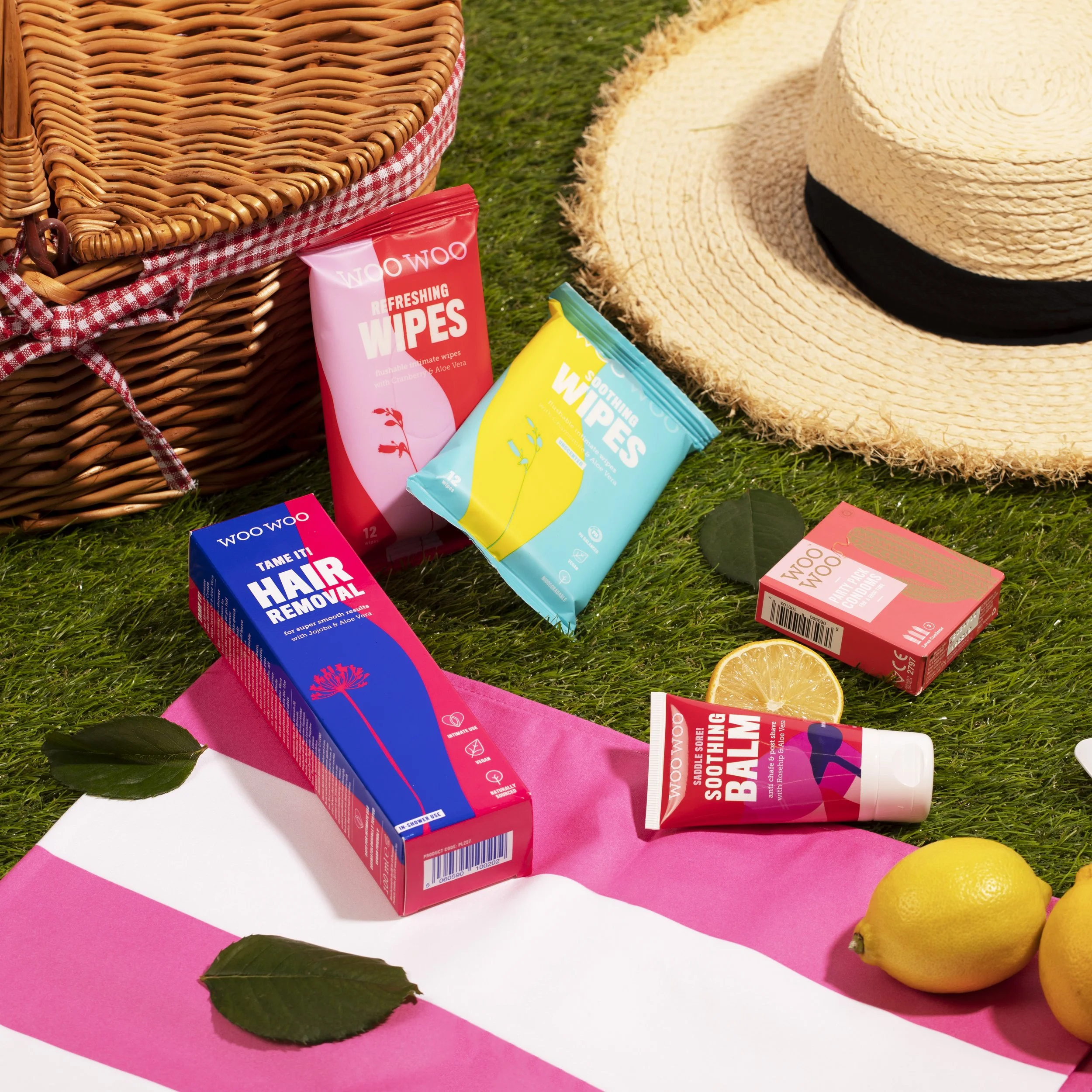 An outdoor picnic scene with a woven basket, a wide-brimmed straw hat, lemon fruits, and various WOO WOO personal care products like refreshing wipes, hair removal, soothing balm, and cooling powder on green grass, with pink and white striped cloth u