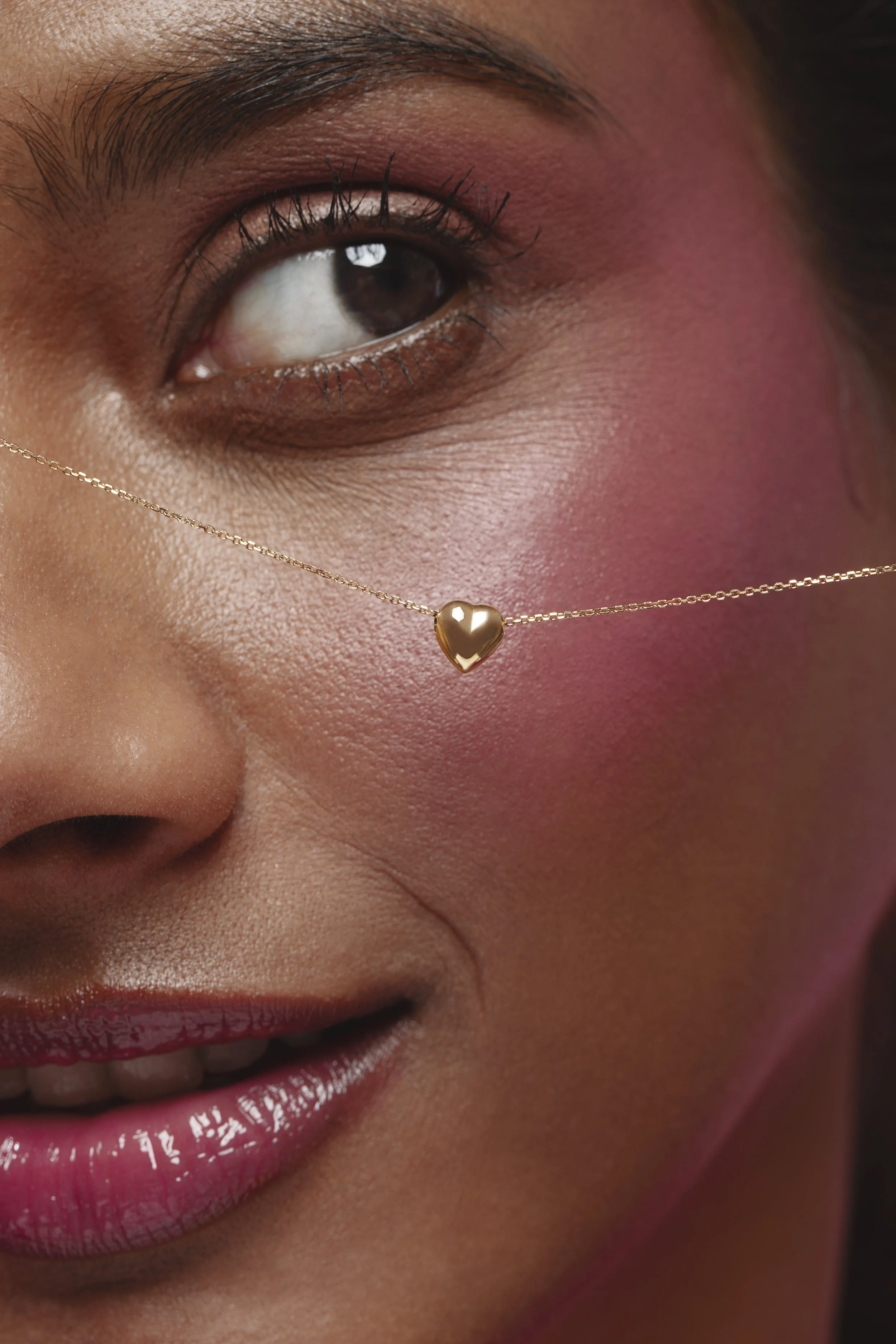 Close-up of a woman wearing a gold heart-shaped necklace, with makeup including pink eyeshadow, mascara, and glossy pink lipstick.
