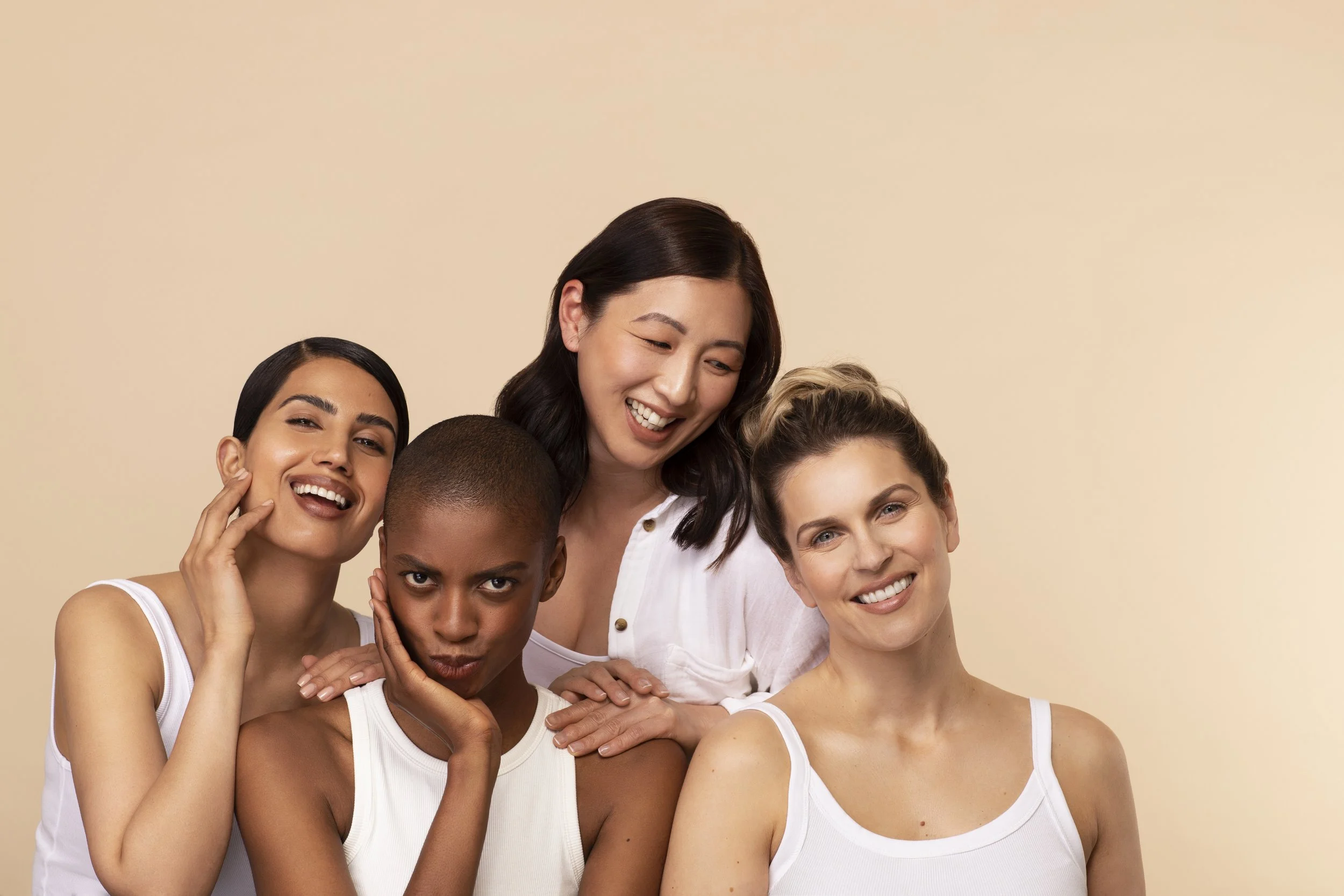 Skincare Group of four diverse women smiling and posing closely together against a beige background.