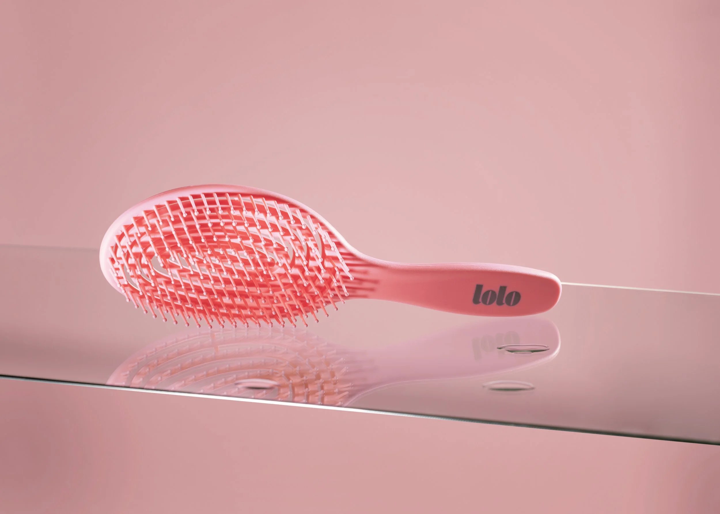 A pink hairbrush with a grid of flexible bristles, labeled 'lolo', resting on a reflective surface with a soft pink background.